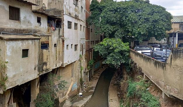 A photo of run down, infomal buildings next to a concrete ravine. There is a tree at the back and a parking garage on the right side.