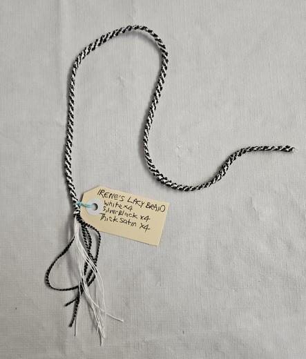 A black and white braid made of eight strands, flat but twisted. An overhand knot at one end spreads out into loose strands.
A label is attached: IRENE'S LACY BRAID, White x 4, Silver-Black x 4, Thick Satin x 4