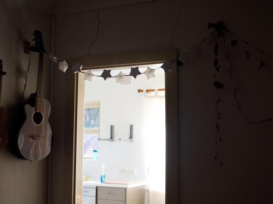 A photograph of a doorway from a dimly lit room, with a string of unlit fairy lights across the doorway, with paper star-shaped "lanterns". A dead pot plant is dimly visible, and so is an acoustic guitar.