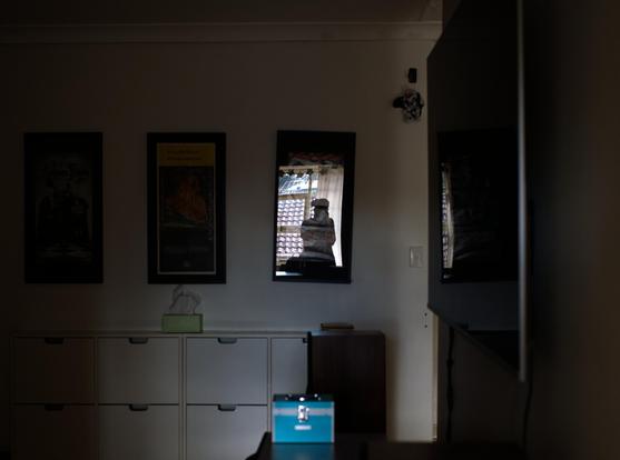 A woman, the photographer, (me) reflected in a crooked poster frame in a dimly lit room of the house she had to abandon when she fled domestic violence