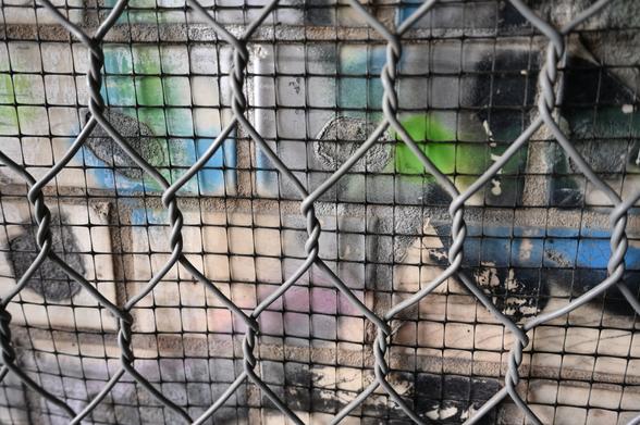 Drahtzaun mit zwei Schichten: im Vordergrund ein grobmaschiger Metallzaun, dahinter ein feinmaschiger schwarzer Gitterdraht. Im Hintergrund ist eine bemalte Wand mit buntem Graffiti in Blau, Grün, Rosa und Schwarz zu sehen. Die Szenerie sieht kaputt und mit Patina belegt aus. Sie stammt aus Oranienburg. 


Wire fence with two layers: in the foreground a coarse metal chain-link fence, behind it a fine black wire mesh. In the background, a painted wall with colorful graffiti in blue, green, pink, and black is visible.