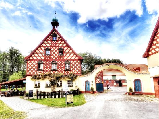 Restaurant in Tschechien, Fachwerkbau, links eine überdachte Terrasse, rechts ein Torbogen, der zu den Pferdeställen führt, der Himmel frühlingshaft weiß blau.