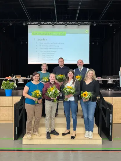 Fotografie von sieben Personen auf einer Bühnentreppe vor einer Leinwand. Die Personen haben Sträuße mit Sonnenblumen in der Hand.