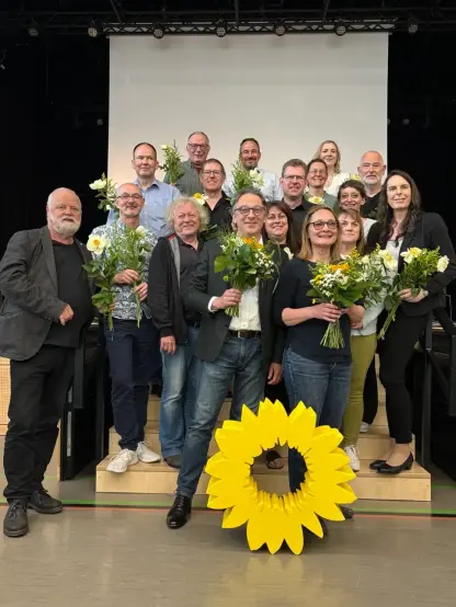 Fotografie von siebzehn Personen auf einer Bühne vor einer Leinwand. Die Personen haben Sträuße mit Sonnenblumen in der Hand.
Vor der Gruppe steht eine große gelbe Sonnenblume