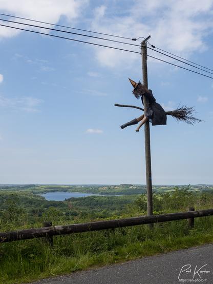 Photo en couleur humoristique de format vertical, présentant une sorcière (un mannequin féminin, vêtue d'un chapeau pointu, d'une robe débraillée, largement entrouverte, et d'une seule botte au pied droit) sur son balai, qui est entrée en collision avec un poteau téléphonique au bord de la route. On a vraiment l'impression d'un choc violent, les jambes et la tête du mannequin étant pratiquement à l'horizontale du côté gauche du poteau, alors que son tronc est resté du côté droit du poteau. On peut imaginer que la botte gauche a été « éjectée » lors du choc avec le poteau.
La route surplombe de quelques dizaines de mètres un paysage de campagne avec un lac entouré de bois, le tout sous un ciel pratiquement sans nuage.