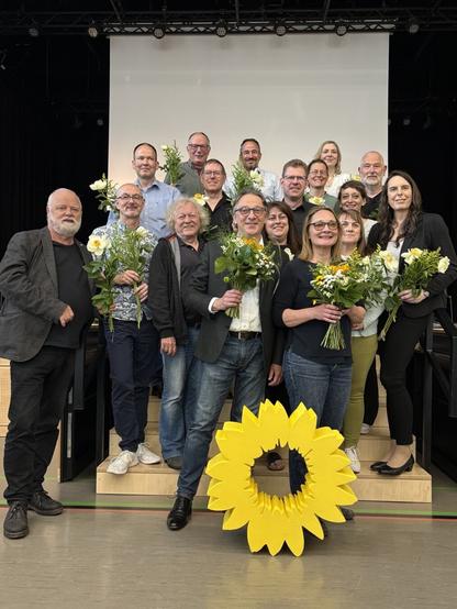 Fotografie von siebzehn Personen auf einer Bühne vor einer Leinwand. Die Personen haben Sträuße mit Sonnenblumen in der Hand.
Vor der Gruppe steht eine große gelbe Sonnenblume