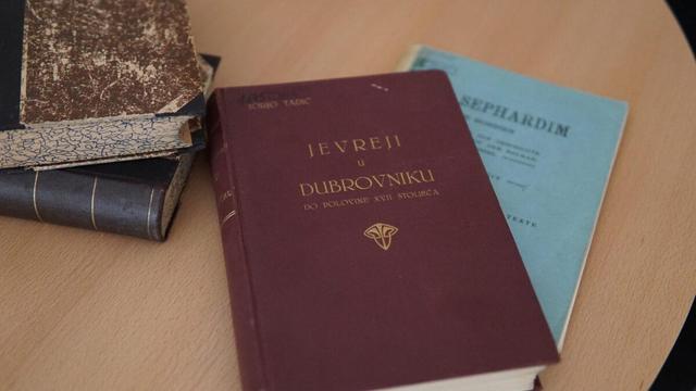 Mehrere historische Bücher auf einem Holztisch, darunter im Vordergrund ein dunkelrotes Buch mit dem Titel "Jevreji u Dubrovniku do polovine XVII stoljeća" (Juden in Dubrovnik bis zur Mitte des 17. Jahrhunderts) von Jorjo Tadić. Dahinter ist ein hellblaues Buch mit dem Titel "Sephardim" zu sehen.