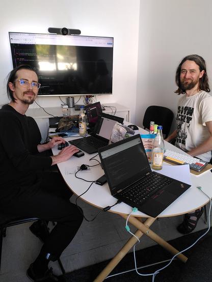 Two people sitting in a smaller room around a table looking into the camera. There's a large monitor attached to the wall showing code. The table is full of laptops, paper, bottles and snacks.