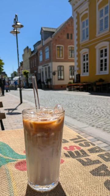 Glas mit geeistem Kaffee auf dem Tisch einer Rösterei, Blick in eine Kopfsteinpflasterstraße mit einem Laternenpfahl und alten Häusern im Hintergrund