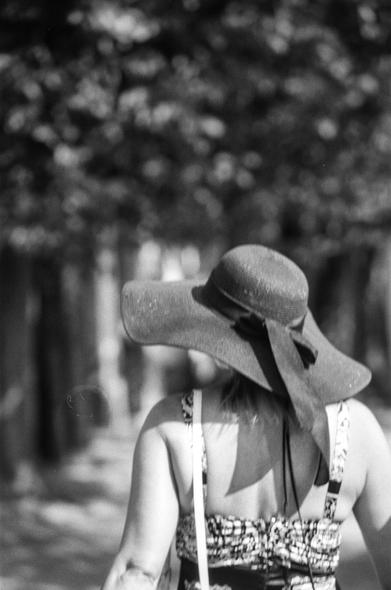 This is a black-and-white photograph taken from behind of a woman walking along a tree-lined path. She wears a large, floppy sunhat tied with a ribbon at the back and a sleeveless, patterned sundress with thin straps. A light-colored shoulder bag strap crosses her back. The background is softly blurred (bokeh), creating a sense of depth and focusing attention on her silhouette and the dappled sunlight filtering through the trees.