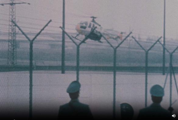 Farbabbildung aus einer Nachrichtensendung. Zu sehen ist ein startender oder landender Hubschrauber hinter einem Zaun, im Vordergrund mehrere uniformierte Polizisten, die den Hubschrauber beobachten.