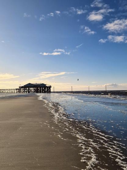 Entlang des Strandes in der Ferne ein Pfahlhaus mit Laufsteg bei Flut. 
Sant Peter Ording