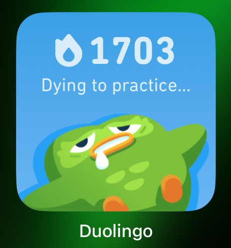 Duolingo app badge that says 1703 and under “dying to practice” with the green owl lying flat on its back, wings splayed out and a smidge of drool coming out of its beak. And apparently it has teeth. As owls do.