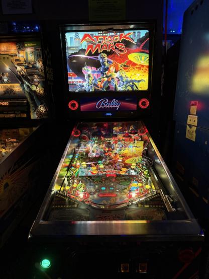 A brightly lit "Attack from Mars" pinball machine in a dim arcade. The vibrant playfield features colorful lights and detailed sci-fi artwork.