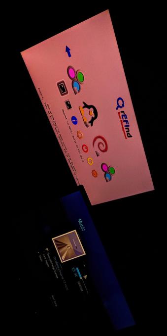 The photograph composed in total darkness shows two screens. The top screen displays a pink background with the text "Q Refind" and a cartoon penguin logo. Below the logo, there are colorful icons, including a spiral, a recycling symbol, and a penguin, along with a blue arrow pointing upwards. The text on the screen reads: "Insert, use arrow keys to move cursor; Enter or F2 for more options; Esc or Backspace to refresh." The bottom screen is dark blue with the word "Music" in light blue text. Below it, there is a music player interface showing the song "Oh Mama Original 12 inch" by Disco & Sussie, with a progress bar indicating 2 minutes and 33 seconds elapsed out of a total duration. The background of the music player is dark, with a small album cover visible.

 Ovis2-8B

🌱 Energy used: 0.232 Wh