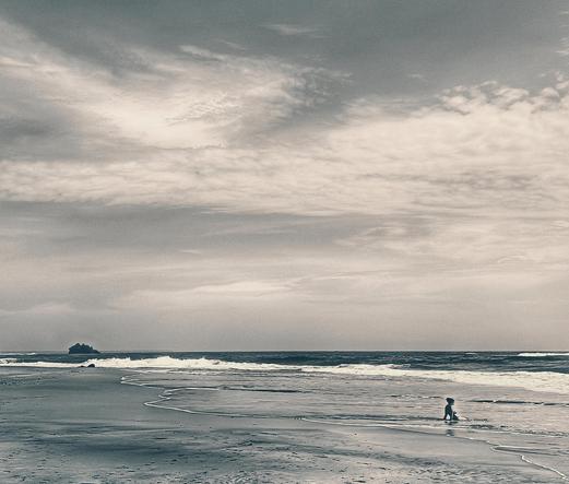 Wide view of Cocles beach in Costa Rica's caribbean. Sepia toned sky and  blue toned beach. A single female figure sits in the sun and enjoys the waves.