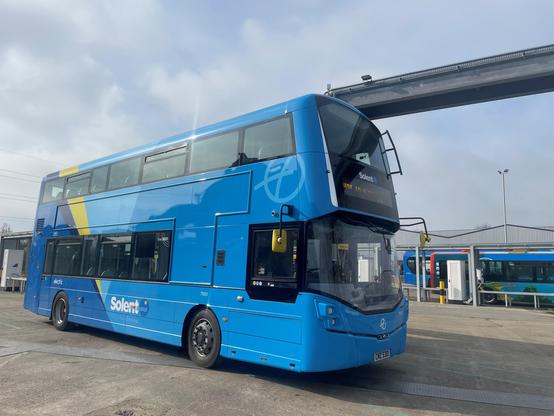 The image shows a blue double-decker bus parked in what appears to be a bus depot. The bus has a contemporary design with large windows on both decks. Its body is painted in a striking blue with a yellow diagonal stripe towards the rear. The word “Solent” and a small logo are visible on the side, while the fleet number “7010” appears near the front. An electronic sign indicates that the vehicle is electric. In the background, another bus with a multicoloured livery and a small building with a canopy can be seen. The scene is set under an overcast sky with soft, diffused light.

Photo by First Bus