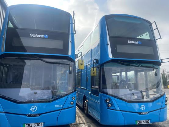 The image shows two modern double-decker buses parked side by side. Both are painted in a vivid blue and feature sleek front designs with large, angled windscreens. The name “Solent” is prominently displayed at the top front of each bus in white text on a dark background, just above the electronic display panels. The bus on the left is marked with fleet number “71004” and registration “CMZ 5304”, while the one on the right is “71002” with plate “CMZ 5302”. They are parked on a tarmac surface under a cloudy sky.

Photo by First Bus