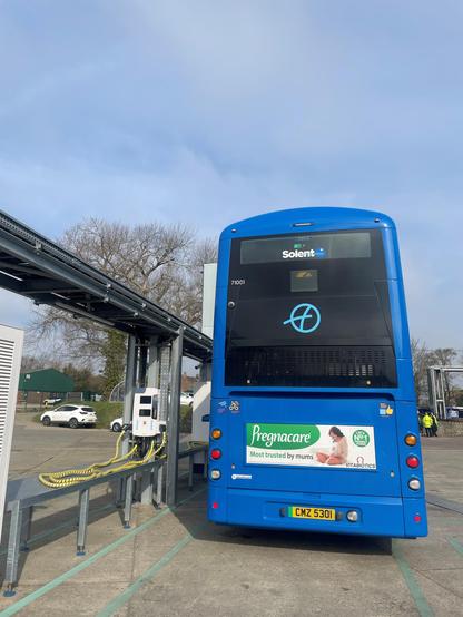 This image shows the rear of a blue electric double-decker bus at a charging station. The bus is connected to a nearby charging unit by yellow cables plugged into the side of the vehicle. In the background are leafless trees and several parked vehicles, including a white car. The sky is overcast with a pale grey hue. The bus has two rear windows and displays several logos and identification numbers. An advert for “Pregnacare”, featuring a seated woman, is displayed on the back. The area is surfaced with concrete and marked with painted lines.

Photo by First Bus