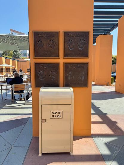 A waste bin labeled "WASTE PLEASE" is positioned in front of a vibrant orange wall displaying four bronze plaques with handprints and names of Disney Legends. The area is sunny, with people seated at tables in the background.