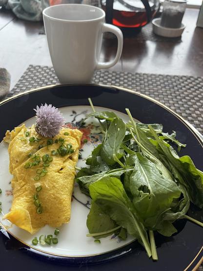 A fancy plate with a folded omelette garnished with chopped chives and flower. It is accompanied by fresh leafy greens. A white cup in the background, along with a glass teapot and infuser