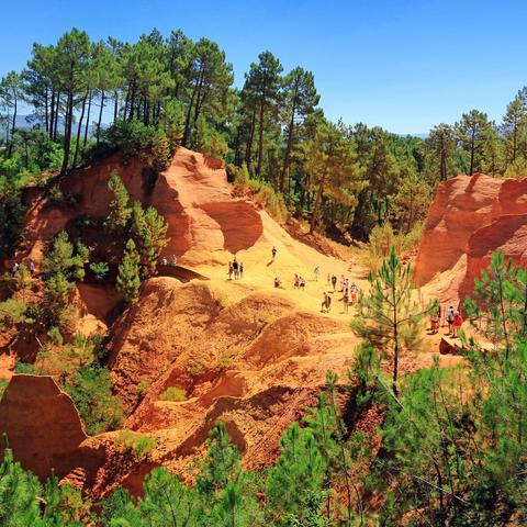 Vibrant orange hills of ochre in layered shades of red, yellow, and orange, dotted with green trees under a bright blue sky.