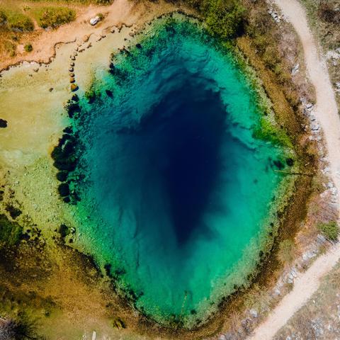 Aerial view of a vibrant deep blue karst spring, its clear waters revealing depth and contrast against the surrounding stony ground and scattered trees.