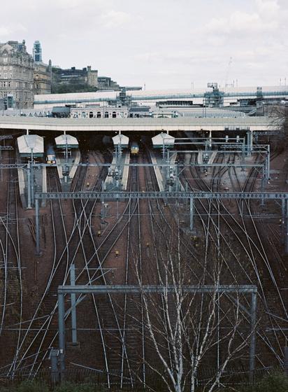 A view of Edinburgh Waverley station from outside the National Gallery of Scotland. Railway tracks rise from the bottom of the image and combine and split before they go into the platforms in the centre and disappear under the canopy.