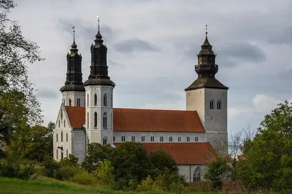 The image depicts a church or a cathedral with two distinct towers, featuring a combination of stone and wooden architectural elements. The structure has a sloped roof and is surrounded by greenery, suggesting a serene, rural setting. The sky appears overcast, adding a moody atmosphere to the scene. The overall design suggests a historical significance, possibly with elements of Gothic or Romanesque architecture.

Image Credits: Wikimedia / Susanne Nilsson / CC BY-SA 4.0