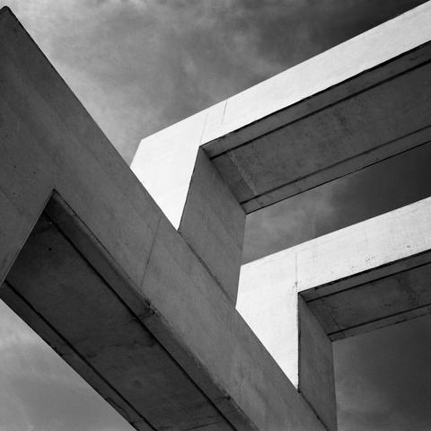 Eine luftige Betonkonstruktion von schräg unten aufgenommen, so dass der Himmel zwischen den Betonstreben hindurch zu sehen ist. Quadratische Schwarz-weiß-Aufnahme mit eher düsterer Stimmung.