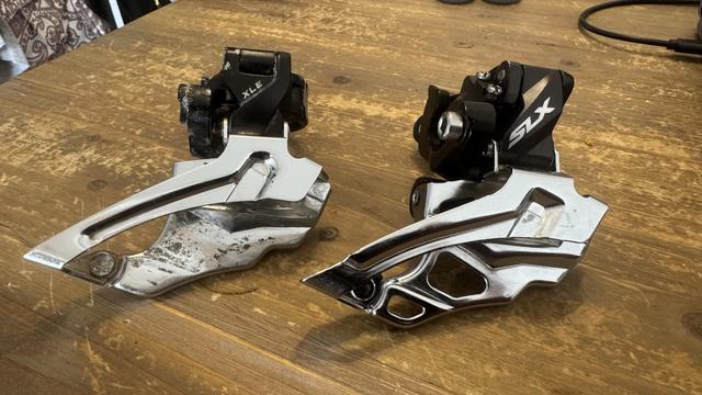 Zwei Fahrrad-Umwerfer sind auf einer Holzoberfläche ausgestellt. Der linke Umwerfer ist mit "XLE" beschriftet, während der rechte mit "SLX" beschriftet ist. Beide haben eine Chromoberfläche mit unterschiedlichen Designs.