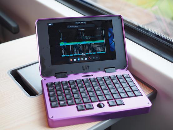 Purple MNT Pocket Reform on a table near the window in a DB Regio Express train. The display shows the Gnome desktop with a terminal running htop.