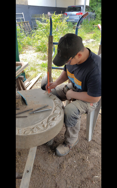 Dieses Foto gibt einen Einblick in unsere "Sommerwerkstatt" - unterm Pavillon in unserem Garten. Unser Geselle sitzt dort vor dem Grabstein, den er gerade fertig arbeitet. Er markiert sich mit einem Bleistift einige Stellen, die er bearbeiten will.

Manchmal ist es wichtig, sich die Arbeit nochmal mit anderem Fokus anzuschauen. 
die Bleistiftlinien helfen dann, dass man die neuen Ideen nicht vergisst, sobald man wieder im Arbeitsmodus ist.