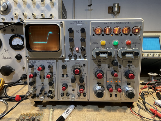 A 1960s oscilloscope with red and black knobs, a blueish square wave trace on the CRT, and a Nixie tube readout