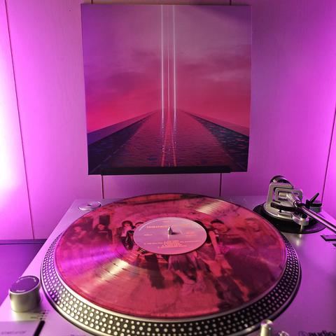 A Pink with Black Smoke vinyl record sits on a turntable. Behind the turntable, a vinyl album outer sleeve is displayed. The front cover shows a long path filled with water, and 3 thin vertical beams of light at the end.