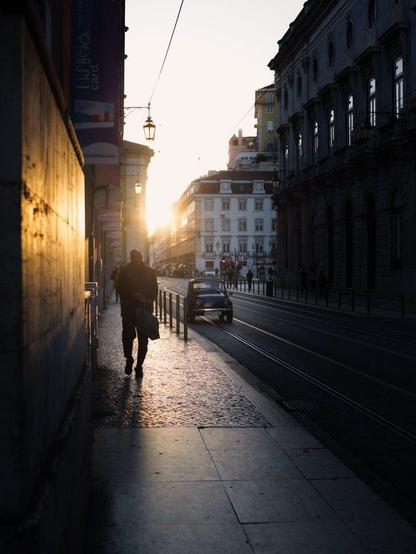 The silhouette of a man walking towards us at sunset, with glaring sunlight on his back coming through the buildings. A classic Fiat 500 is driving towards us as well on the street, parallel to tramway tracks.