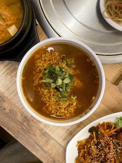 An image of ramyeon, japchae, sundubu-jjigae, and jeyuk-deopbap on a wooden table at a Korean restaurant.