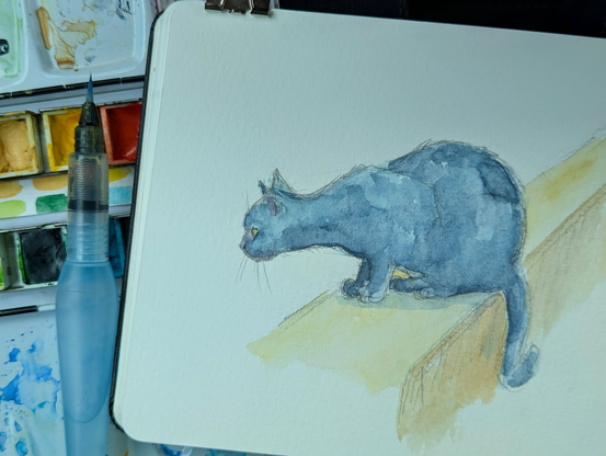 Watercolors, paintbrush and sketchbook. On the sketchbook page a watercolor sketch of a black cat sitting on a wall looking curiously to the left side. Painting by Karen Kaspar