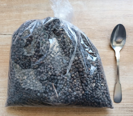 ein großer Plastikbeutel mit ganzem schwarzen Pfeffer. Zum Größenvergleich liegt ein kleiner Löffel daneben.