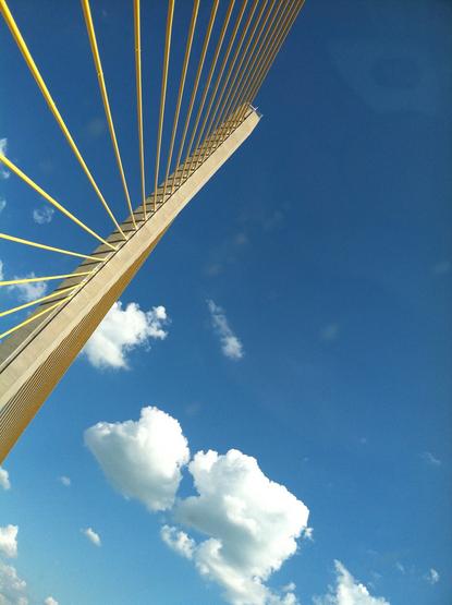 A view of the tower of a yellow suspension bridge from beneath it looking up at a blue sky with a handful of non-threatening clouds.
