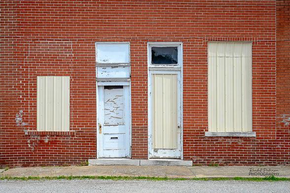 Two doors and two windows in brick building adjacent to a sidewalk and street.  Both windows and one door are covered by cream colored metal roofing material.