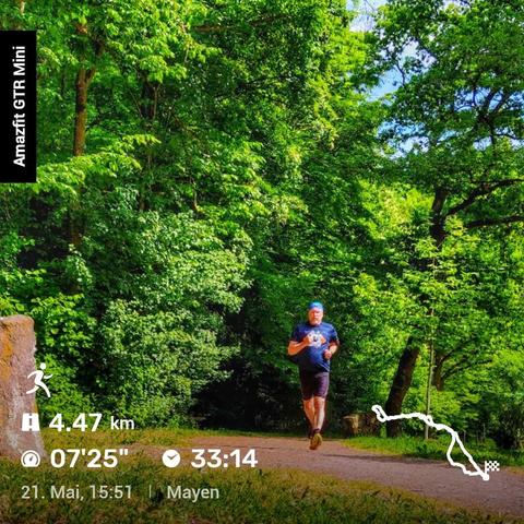 Eine Person joggt auf einem Waldweg, umgeben von dichtem, grünen Laub. Links im Bild liegt ein Stein, während im Hintergrund hohe Bäume die Szenerie prägen. Auf der linken Seite steht der Schriftzug „Amazfit GTR Mini“. Unten im Bild sind Laufdaten zu sehen: Die zurückgelegte Strecke beträgt 4,47 km, mit einer durchschnittlichen Pace von 07'25" pro Kilometer und einer Gesamtzeit von 33:14 Minuten. Das Datum und die Uhrzeit des Laufs sind der 21. Mai um 15:51 Uhr, der Ort ist Mayen. Rechts unten zeigt eine Karte die Laufstrecke.