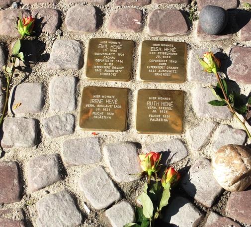 The image shows a series of brass memorial plaques set into a cobblestone path. Each plaque commemorates members of the Hené family, detailing their names, birth years, deportation, and fates during the Holocaust.