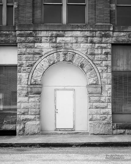 A simple, solid, white entrance door is framed by a large, arched, stone facade on an old building.