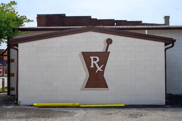 A large brown Rx symbol is painted on the cream-colored wall of a drug store.
