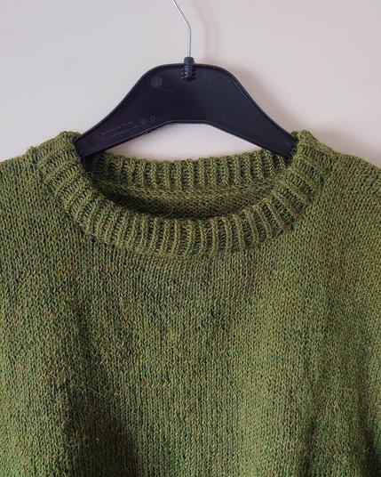 a close up photo of a handknit sweater, focusing mostly on the neckline, which is a folded over collar of 1x1 half twisted rib stitch. the yarn is a warm green with flecks of yellowy brown fibres heathered in
