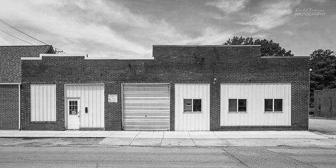 A one story brick commerical building with multiple windows and doors that have been filled in with white metal roofing material.  There are three windows, one door, and a garage door on the face of the building.