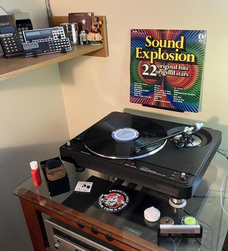 A colorful 1973 K-tel compilation LP titled Sound Explosion: 22 Original Hits Original Stars is playing on a black turntable. The album cover is propped vertically behind the turntable, displaying its iconic rainbow burst design. A Sharp , R2-D2 figurine, and Hawaiian hula dancer sit on a shelf above. Below the turntable are record cleaning supplies.