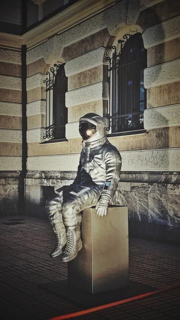 The image displays a full-body sculpture of an astronaut, rendered in a metallic silver material, sitting on a dark, rectangular pedestal. The astronaut is wearing a helmet with a reflective visor that obscures the face. The suit has visible details such as creases, stitching, and some darker blue accents on the chest and arms.
The sculpture is positioned outdoors on a paved area, with a building in the background. The building features light-colored block work and has multiple windows with dark, ornate bars. The windows have rounded arches at the top. The night scene is illuminated by artificial light casting shadows on the ground and parts of the building. A faint red line is visible at the very bottom of the frame.