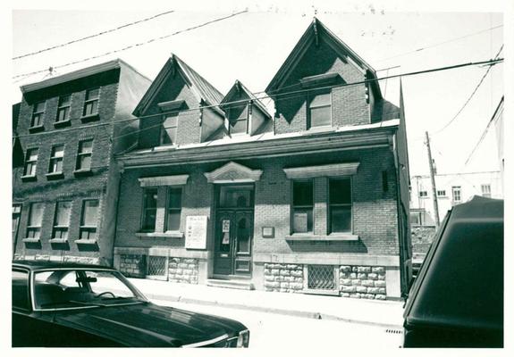 Une photo en noir et blanc d'une maison en brique au toit en pente comportant trois lucarnes: deux grandes de chaque côté, et une petite au centre. La porte d'entrée est située en plein milieu, et est surmontée d'un linteau triangulaire et blanc. Sur le côté gauche de la porte on voit un panneau d'affichage, on ne distingue pas ce qui y est écrit.  De part et d'autre de la porte des fenêtres doubles. Le solage semble fait de pierres taillées.  Du côté gauche de la maison, un édifice en brique à trois étages. Des fils électriques passent dans le champs de la photo. Au bas de la photo, une automobile qui date sans doute de la fin des années '70.
